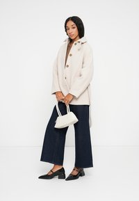 Crème-kleurige oversized jas met grote knopen, gecombineerd met een bruin gebreid topje, lichte-wijd broek en een kleine witte handtas. Zwarte hakken.