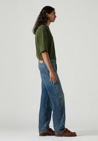 Green oversized T-shirt, blue straight-leg jeans, and brown leather shoes. Notable details include a side pocket and a leather patch on jeans.