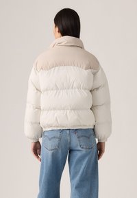 Lett puffer-jakke med en hvit og beige fargeblokkdesign, myk tekstur, høy krage og elastiske mansjetter og kant.