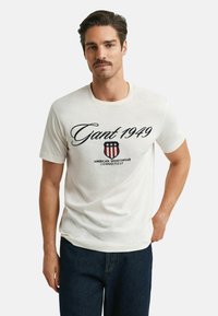 Hvid bomulds t-shirt med sort tekst, der står "Gant 1949", samt et rødt, hvidt og blåt skjoldlogo; rund hals og korte ærmer.