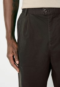 Black trousers made of smooth fabric featuring a pleated front, a button closure, and side pockets. Hand resting on the waistband.