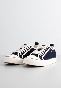 Baskets en toile bleu marine avec semelles en caoutchouc blanc, dotées de lacets crème, de panneaux latéraux contrastants et d'un petit logo sur le côté.