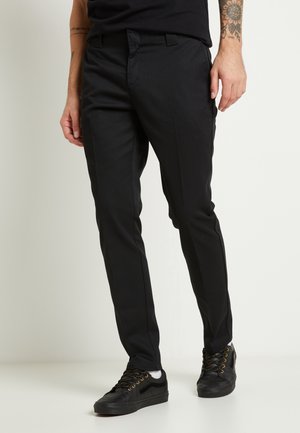 Man draagt zwarte slim-fit broek, zwarte sneakers met gouden veteroogjes en een zwart shirt, staand tegen een effen lichte achtergrond.
