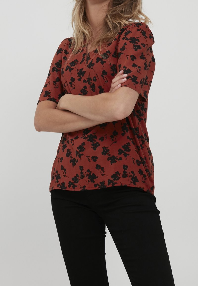 Blouse couleur rouille avec un motif floral noir, à manches courtes, coupe décontractée et texture de tissu douce. Assortie à un pantalon noir.