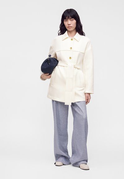 Femme aux longs cheveux foncés porte un manteau blanc ceinturé, un pantalon large gris clair, des chaussures beiges et tient une pochette tricotée bleu marine.