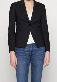 Černý šitý blazer s klopami, zapínáním na jeden knoflík a dvěma předními kapsami. Nosený přes bílé tričko a kombinovaný s modrými džíny.