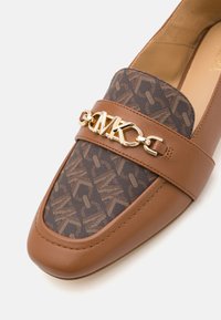 Brun läder loafer med mönstrad tygfront, guldkedjedetalj med "MK" initialer ovanpå.