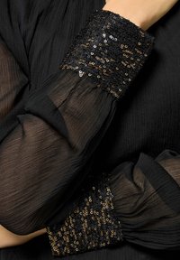 Blusa nera in tessuto trasparente con texture plissettata. Maniche adornate con paillettes nere e oro. Design elegante con maniche ampie.