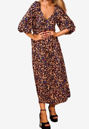 Vrouw draagt een midi-jurk met luipaardprint en pofmouwen, gecombineerd met zwarte platform sandalen met hoge hakken, handen rustend op de taille.