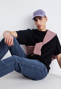 Zwarte t-shirt, lichtpaarse pet met klein logo, en lichtroze sweater over de schouders gedrapeerd. Blauwe jeans met verticale stikdetails.