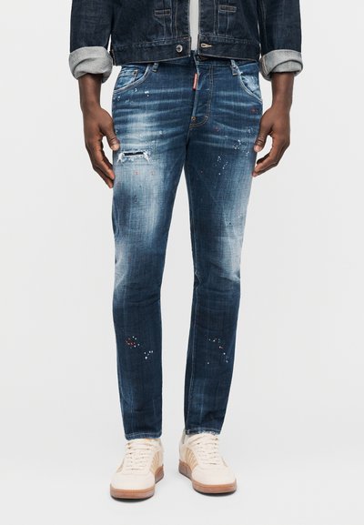 Jeans blu scuro strappati con schizzi di vernice indossati con giacca di jeans risvoltata e sneaker beige su sfondo chiaro.