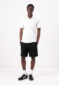 Weißes Poloshirt mit kurzen Ärmeln und Tasche, kombiniert mit schwarzen knielangen Shorts und schwarzen Sneakers mit weißen Akzenten, vor einem neutralen Hintergrund stehend.