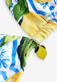 Bunter Stoff mit einem Zitronen- und Blumenmuster, versehen mit einem goldenen Zitronenanhänger am geschwungenen Rand. Sanfte Textur.