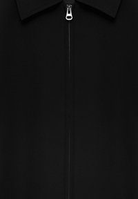 Vêtement noir avec col et fermeture éclair centrale argentée, fermeture éclair partiellement fermée, montrant le détail de la couture le long de la ligne de la fermeture éclair.