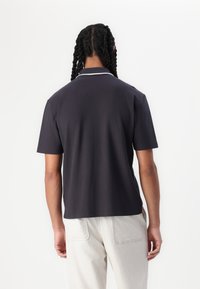 Cămașă polo neagră cu mânecă scurtă și guler clasic, cu cusături de accent albe, purtată cu pantaloni texturați de culoare deschisă.