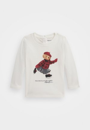 T-shirt bianco a maniche lunghe con una grafica di un orso che indossa una camicia a quadri rossi, una gonna di jeans e scarpe nere, con il testo "Polo Bear by Ralph Lauren".
