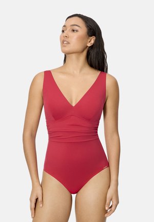 Mujer con cabello largo y oscuro, vestida con un bañador rojo de una pieza sin mangas, mirando ligeramente hacia su izquierda sobre un fondo liso.