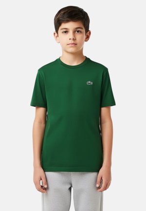 Ragazzo che indossa una maglietta a maniche corte verde scuro con un piccolo logo di un coccodrillo sul petto e pantaloni grigio chiaro, in piedi con il volto rivolto in avanti.