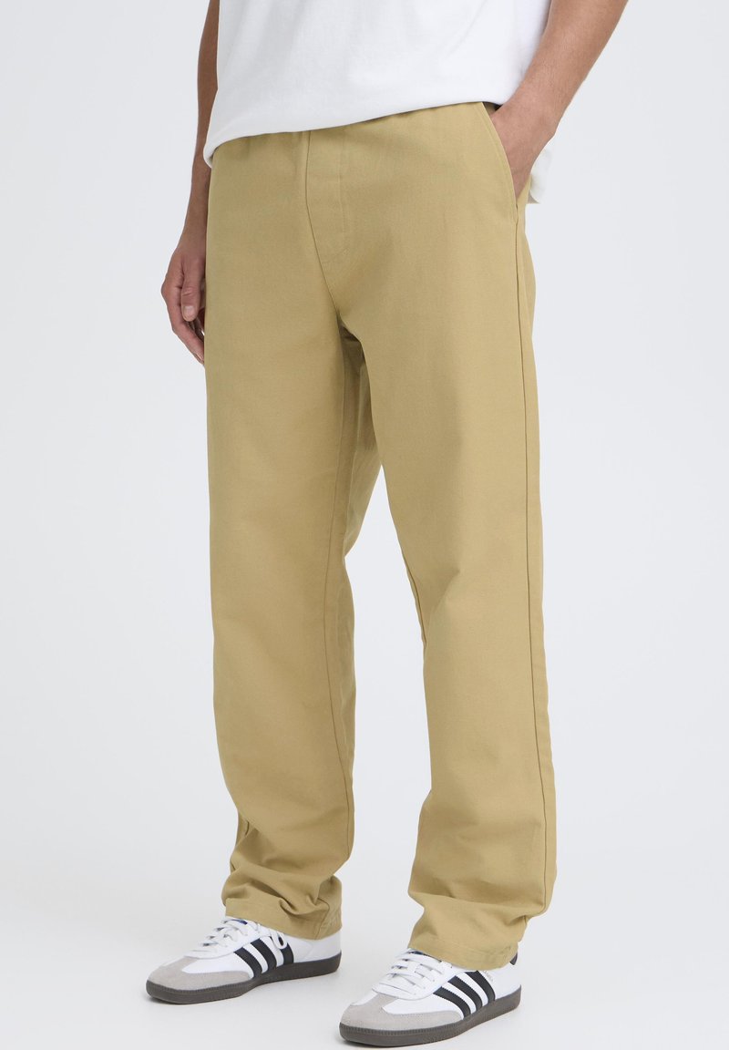 Pantaloni beige a gamba dritta con una texture liscia, dotati di tasche laterali e una vestibilità comoda. Abbinati a scarpe sportive nere e bianche.