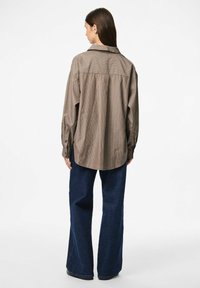 Rjavo-bež prugasta srajca z gumbi, ovratnikom, dolgimi rokavi in sproščenim krojem, v kombinaciji z temno modrimi širokimi hlačami.