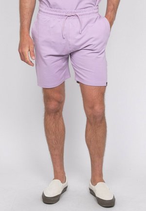 Homme portant un short à cordon violet clair et un t-shirt assorti, les mains dans les poches, et des chaussures blanches sans lacets sur fond uni.