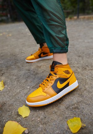 Żółto-czarne wysokie trampki Nike Air Jordan noszone z zielonymi spodniami na ziemnej ścieżce z rozrzuconymi żółtymi liśćmi.