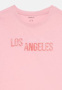 Rozā kokvilnas t-krekls ar uzrakstu "LOS ANGELES" treknām, spīdīgām sarkanām burtiem un apakšā smalkāk rakstītu "Athletic Division" baltajā krāsā.