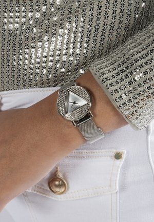 Zápästie s hodinkami so strieborným sieťovým remienkom, trojuholníkovým ciferníkom a kryštálmi, doplnené strieborným flitrovaným dlhým rukávom a bielymi nohavicami s gombíkovými vreckami.