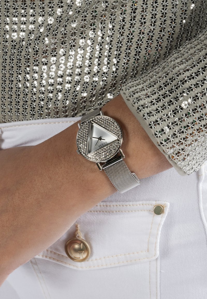 Ruka s satom sa srebrnom mrežastom narukvicom, trokutastim brojčanikom i kristalima, uparenim sa srebrnom bluzom dugih rukava ukrašenom šljokicama i bijelim hlačama s gumbima na džepu.