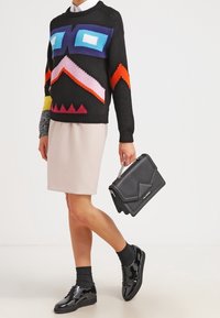 Schwarzer gestrickter Pullover mit bunten geometrischen Mustern, beiger Rock, glänzende schwarze Schuhe und eine schwarze Handtasche mit silberner Kettenriemen.