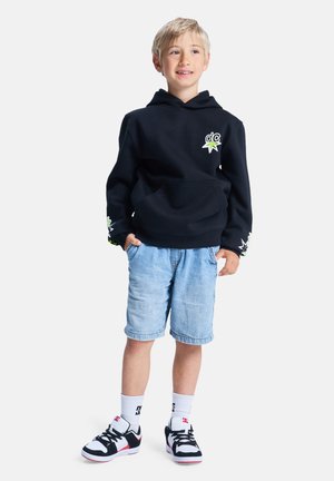 Sweat à capuche noir avec une poche avant et un logo, short en jean bleu clair, chaussettes blanches, et baskets noir et blanc avec des accents rouges.