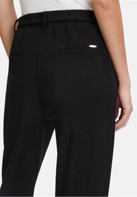 Zwarte broek met een gladde, gestructureerde stof, elastische tailleband en twee achterzakken met subtiele metallic accenten.