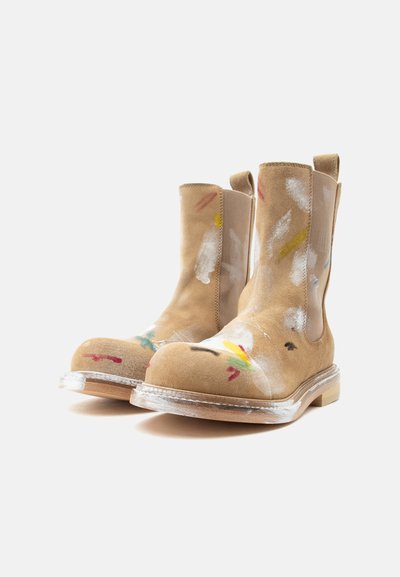 Τακούνια μποτάκια σε ταμπά σουέτ με θηλύκια, διάφανα πέλματα και πολύχρωμες πιτσιλιές βαφής στα δάχτυλα και τους άξονες, τοποθετημένα σε λευκό φόντο.