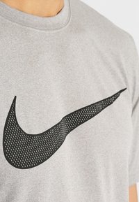 Grå bomullströja med en svart perforerad Nike swoosh-logotyp som är tydligt placerad på framsidan. Rundad halsringning, korta ärmar.