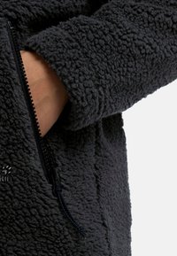 Zwarte fleece jas met een gestructureerd, fuzzy oppervlak. Inclusief een ritszak met een zwarte rits en een lusje.