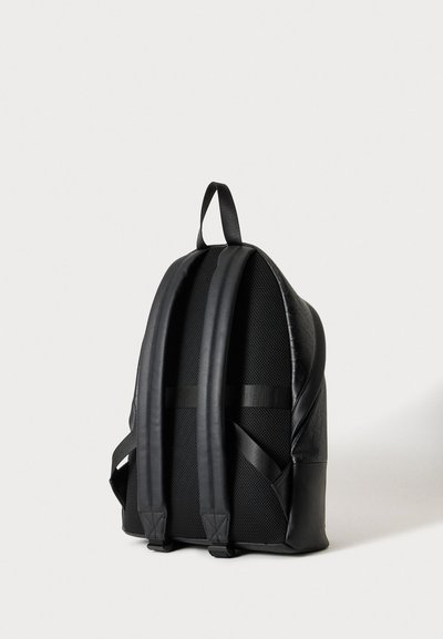 Sac à dos noir vu de dos, avec des bretelles rembourrées en maille, une poignée de transport sur le dessus et une poche latérale sur un fond clair uni.