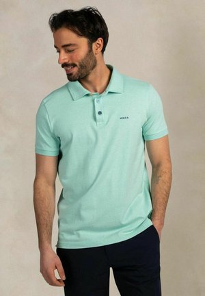 Man met donker haar en baard, draagt een lichtblauw poloshirt en donkere broek, kijkt naar beneden met één hand in de zak.