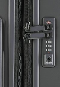Esquina de una maleta rígida gris con cremalleras negras, tiradores de cremallera plateados dobles y un candado de combinación con el insignia de la TSA y número de identificación.