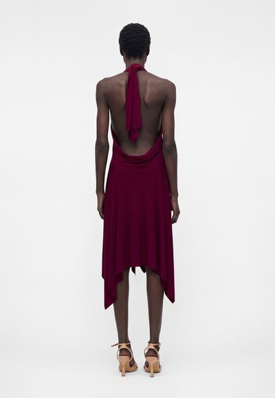 Γυναίκα όρθια με πλάτη, φοράει αμάνικο, ανοιχτή πλάτη σκούρο κόκκινο φόρεμα με μπεζ ψηλοτάκουνα παπούτσια με λουράκι στον αστράγαλο σε απλό λευκό φόντο.