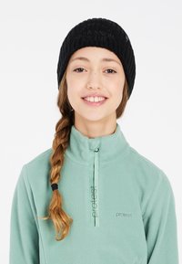 Cappellino di maglia a coste nera con un motivo testurizzato, abbinato a un top in fleece verde chiaro con chiusura a mezza zip e dettaglio del logo.