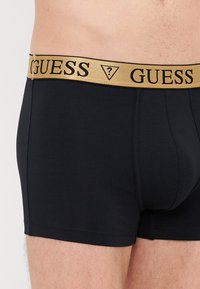 Melnas bokseru bikses ar zelta elastīgo jostu, uz kuras rakstīts "GUESS". Viegla, gluda materiāla, pieguļošs dizains, bez redzamām rakstu vai akcentu detaļām.