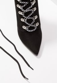 Black suede high-heeled boot with pointed toe, silver hardware, and black and white checkered laces, accompanied by a separate black lace.