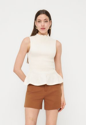 Γυναίκα με μακριά καστανά μαλλιά που φορά χρυσούς κρίκους στα αυτιά, αμάνικη λευκή μπλούζα με πεπλούμ και καφέ σορτς, στέκεται με το χέρι στην τσέπη.