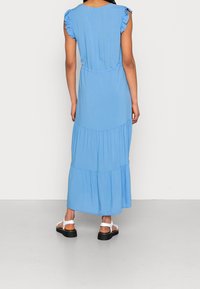 Vaaleansininen maksimekko, jossa on rypytetyt hihat, istuva vyötärö ja kerroksellinen helma. Pehmeä kangas; nilkkapituisuus. Yhdistetty paksupohjaisiin mustiin sandaaleihin.