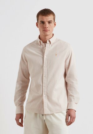 Jonge man met kort haar, gekleed in een lichtroze, langermelig overhemd met dunne blauwe strepen en een witte broek, staand tegen een witte achtergrond.