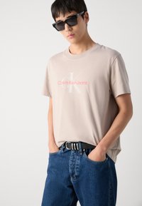T-shirt en coton beige avec un logo "Calvin Klein Jeans" en blanc et rose, associé à un jean en denim bleu et une ceinture noire avec des accents argentés.