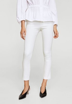 Kobieta ubrana w dopasowane białe spodnie do kostek, białą bluzkę z elastyczną talią i czarne czółenka na obcasie z odkrytą piętą, stojąca na jednolitym tle.