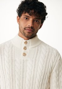 Pull crème en tricot torsadé avec un col montant et trois boutons en bois. Motif texturé dans un tissu épais et douillet.