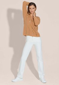 Bruine geribbelde trui met zigzagpatroon en lichte accenten, gecombineerd met witte nauwsluitende jeans en witte sneakers. Neutrale achtergrond.