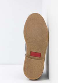 Semelle de chaussure avec une bande de roulement en caoutchouc texturé marron clair et un logo rectangulaire rouge Kickers près de la zone de la voûte plantaire, positionnée verticalement sur un fond blanc.